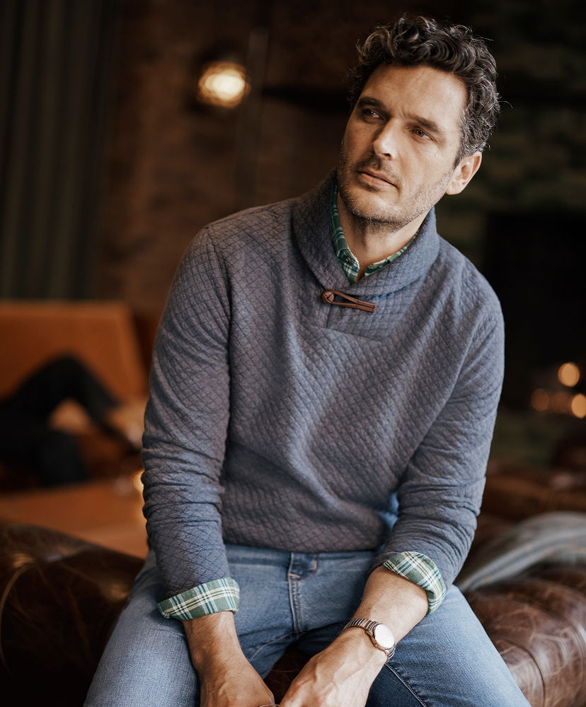Man sitting on a couch, wearing a sweater and looking thoughtfully.