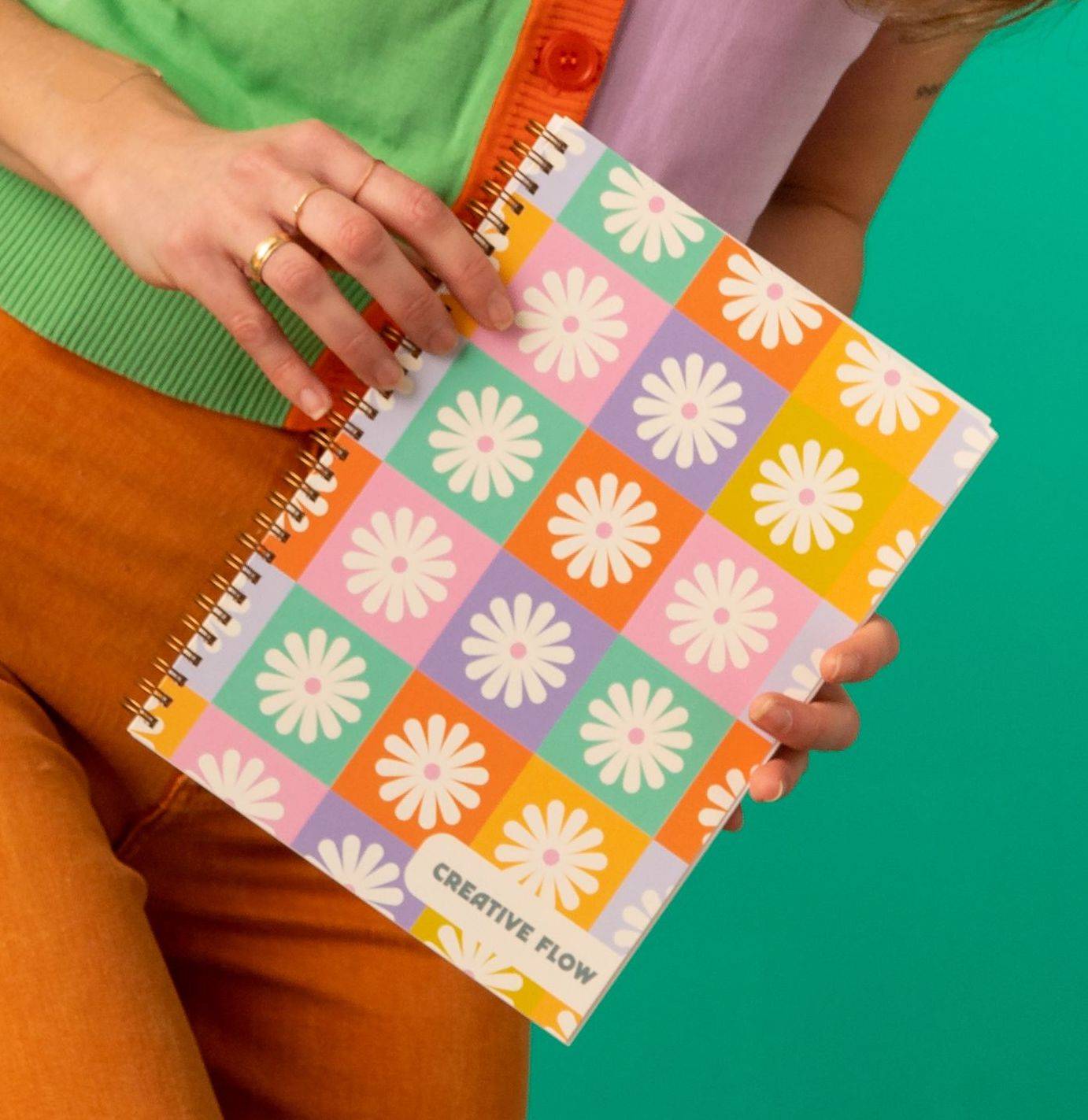 A close-up shot of a woman holding the Creative Flow Journal. It is a journal with space to doodle your day and express your creativity! The cover has a colorful grid pattern with white daisy's in each square. 