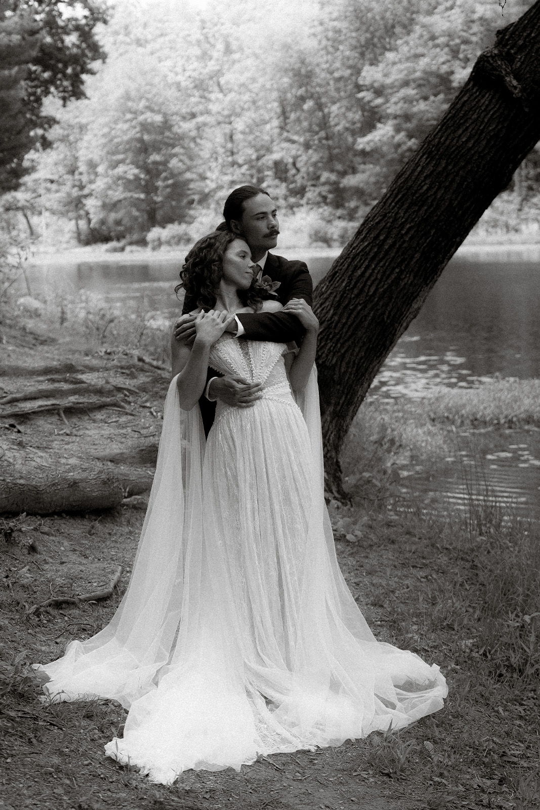 A couple embraces by a serene lake in black and white.