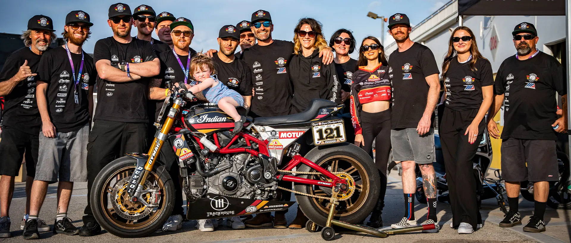 A group photo of the Roland Sands Design racing team at a motorcycle racing event. The team, wearing matching black T-shirts adorned with sponsor logos, gathers around a red and black Indian motorcycle. A young child sits on the motorcycle, surrounded by smiling team members in a celebratory atmosphere.