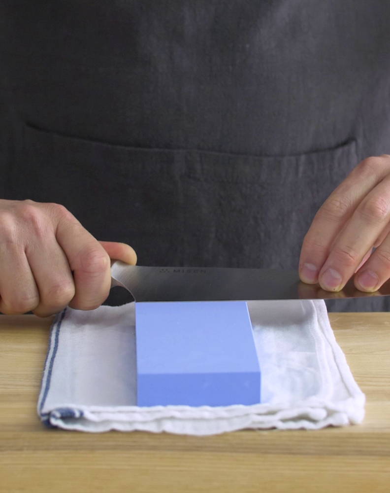 A chef sharpening a Misen Chef’s Knife on a blue 1000-grit Misen Sharpening Stone placed on a kitchen towel.