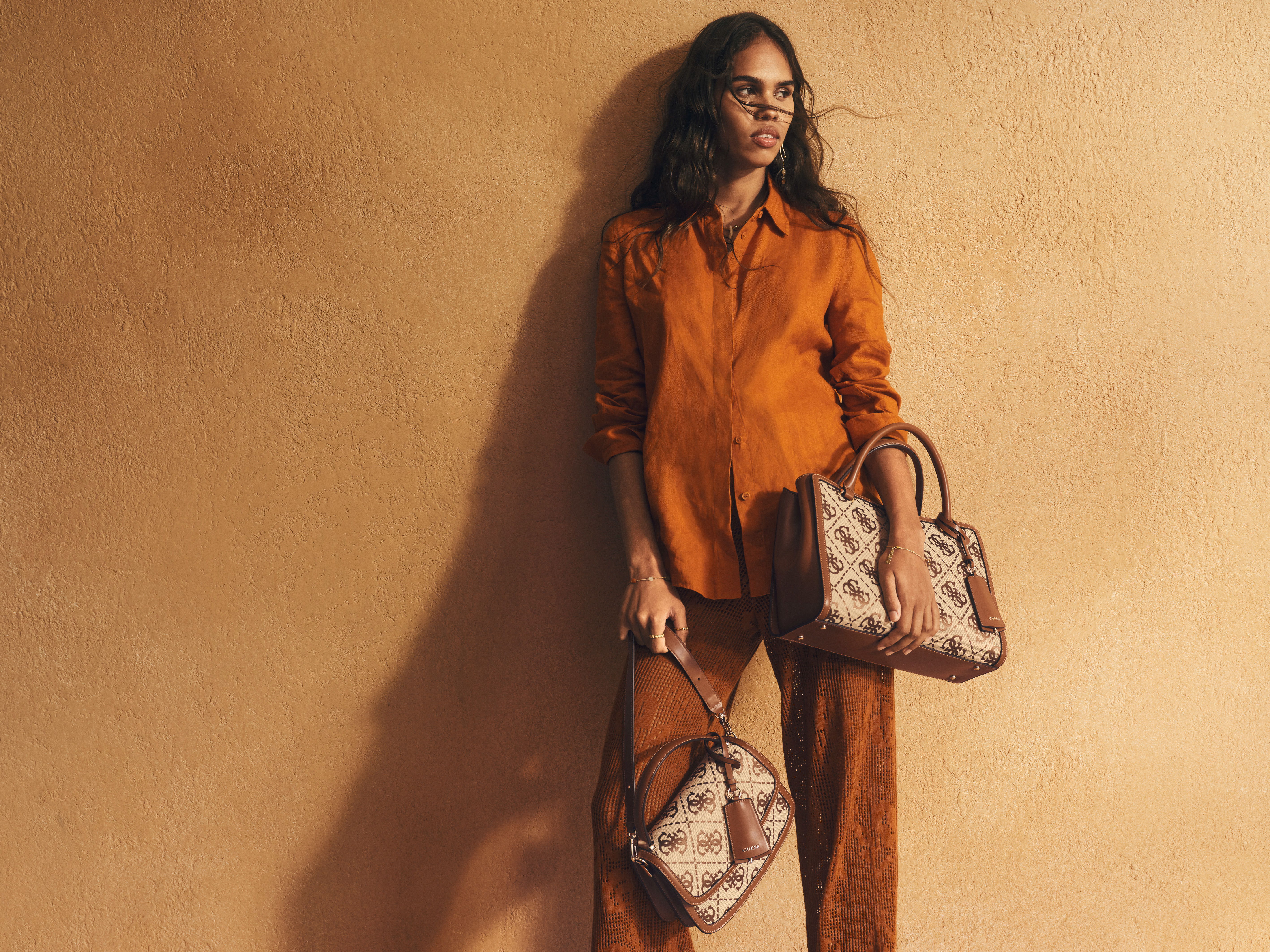 Woman in orange outfit holding two stylish handbags against a warm background.