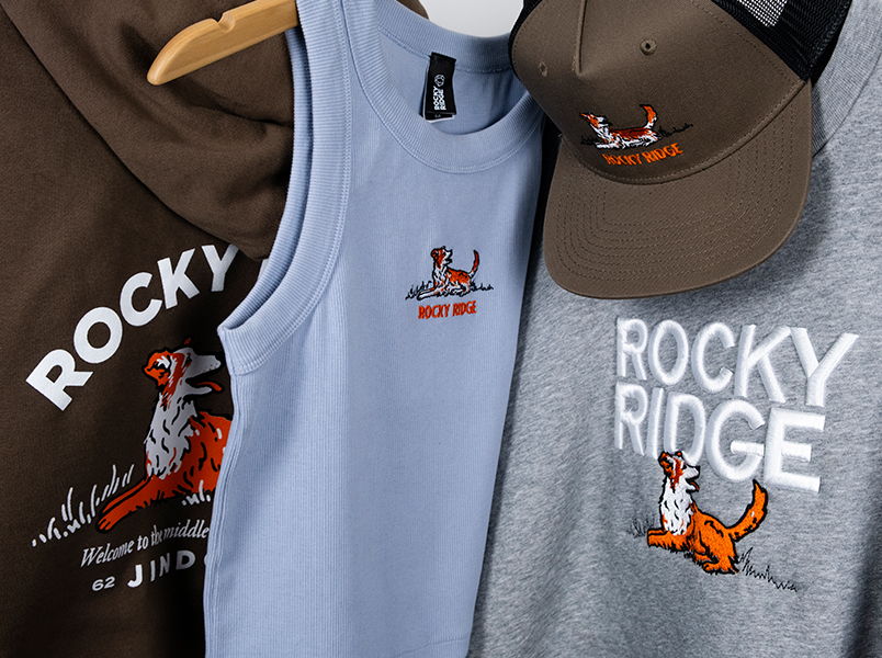 Collection of Embroidered Rocky Ridge Merch featuring Ace the Border Collie on a crop rib singlet, contrast trucker cap and grey marle crewneck