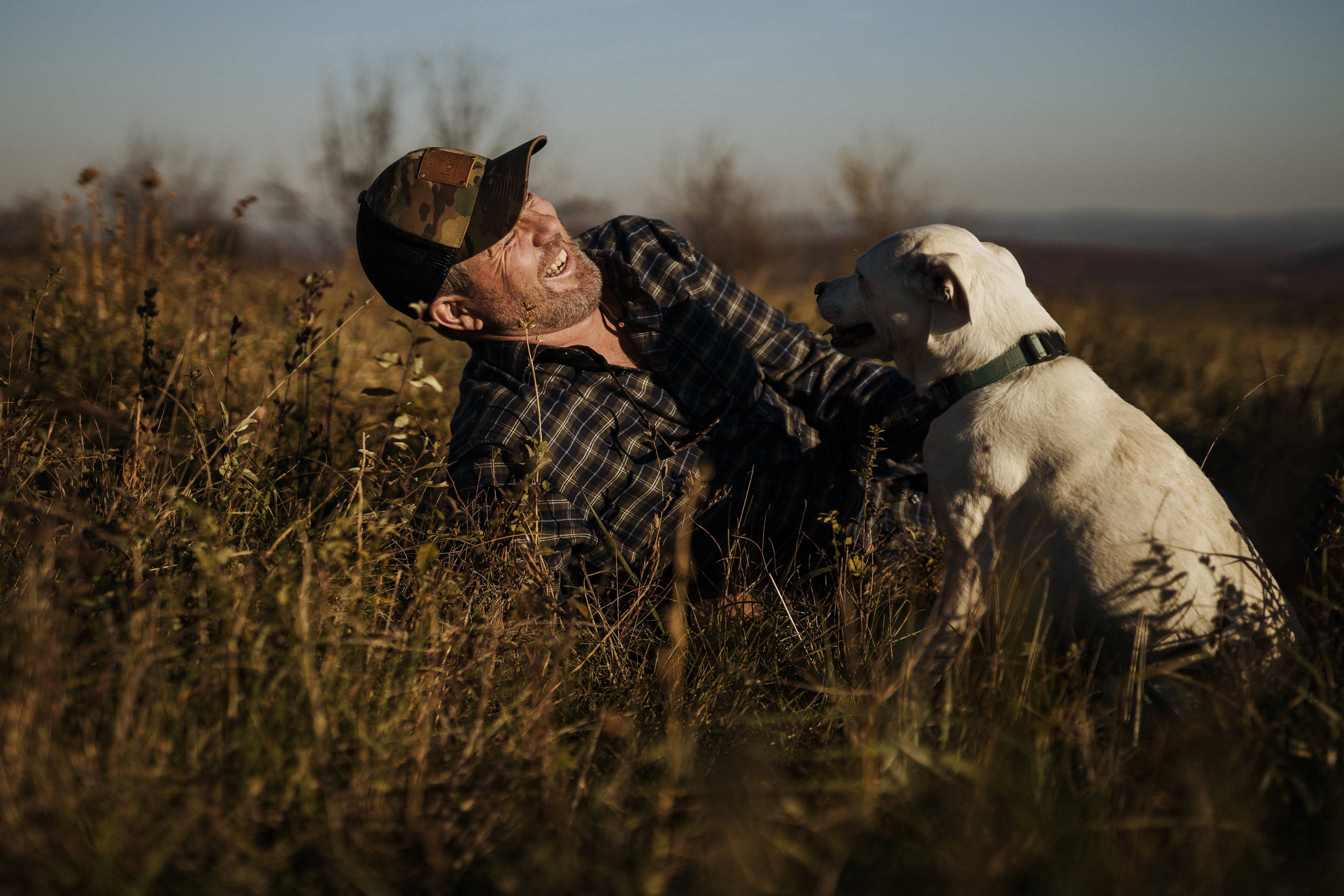 A man laying in a grassy field wearing the Forest Arrowood Flannel custom shirt smiling at his dog.