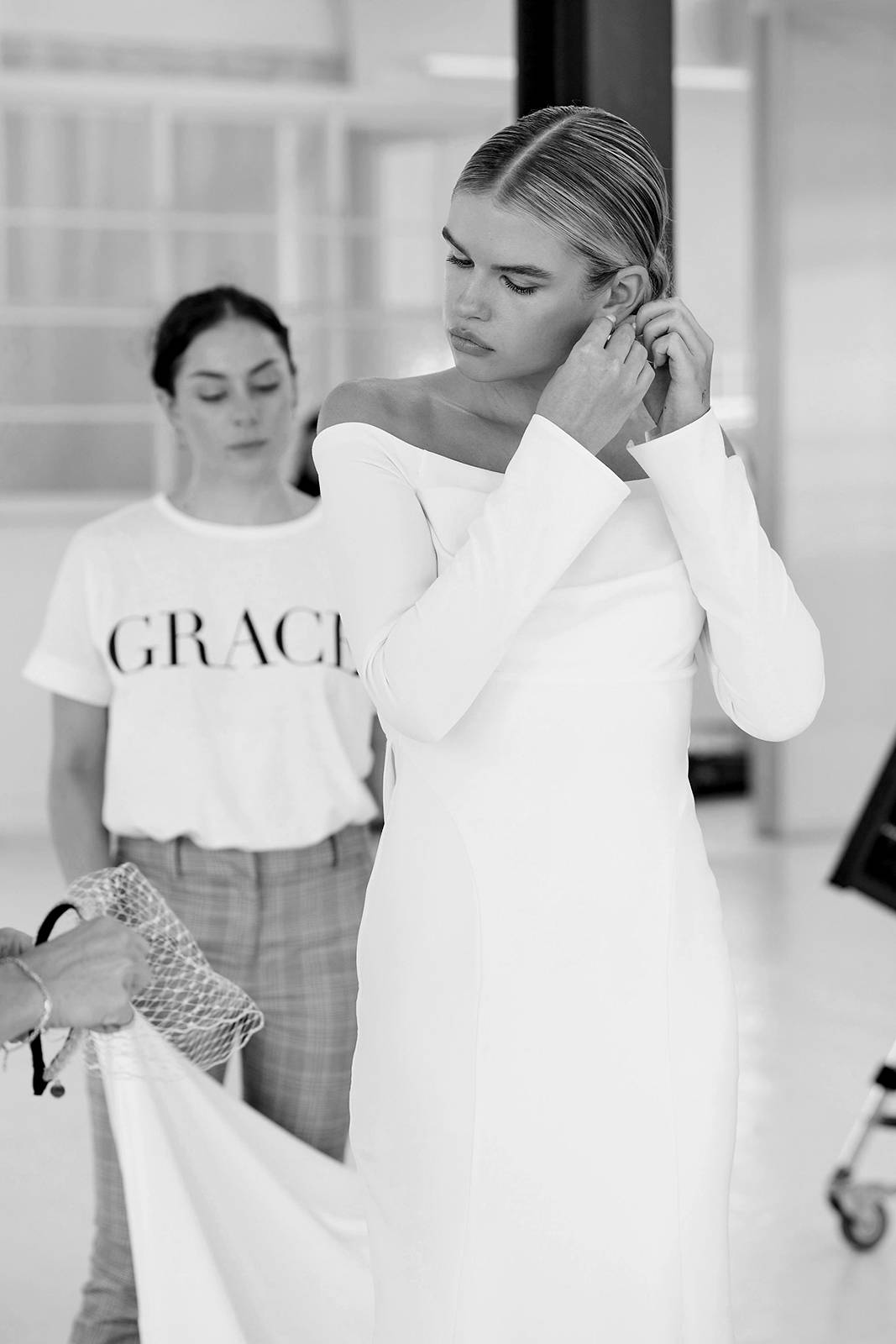Black and white image of model wearing Deia putting her earrings in and stylists adjusting her skirt