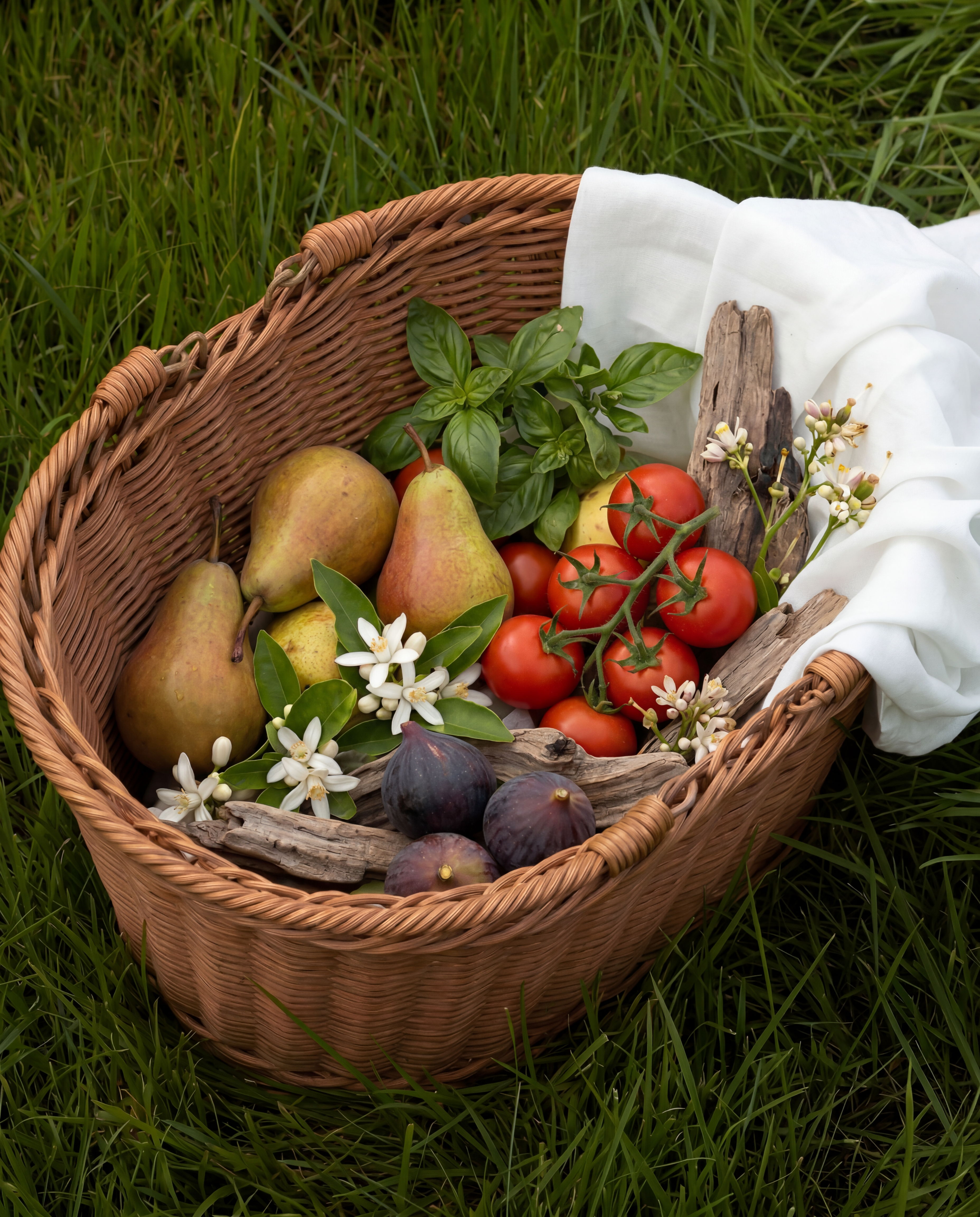 A wicker basket filled with fruits, herbs, and flowers on green grass.