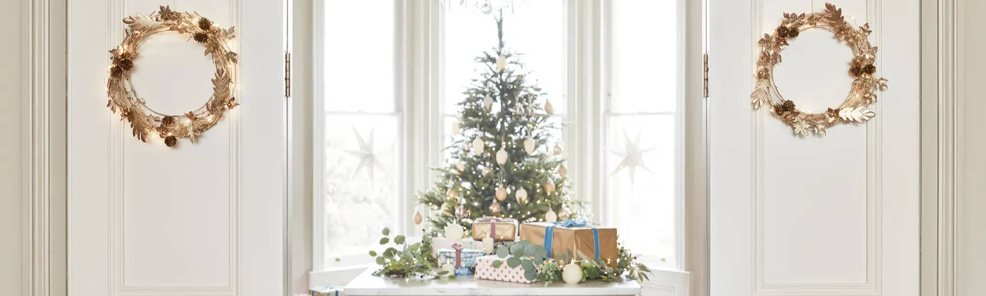 Mit goldenen Kränzen  dekorierte, geöffnete Flügeltür, dahinter ein geschmückter Weihnachtsbaum und auf einem Tisch platzierte Geschenke und TruGlow LED Kerzen
