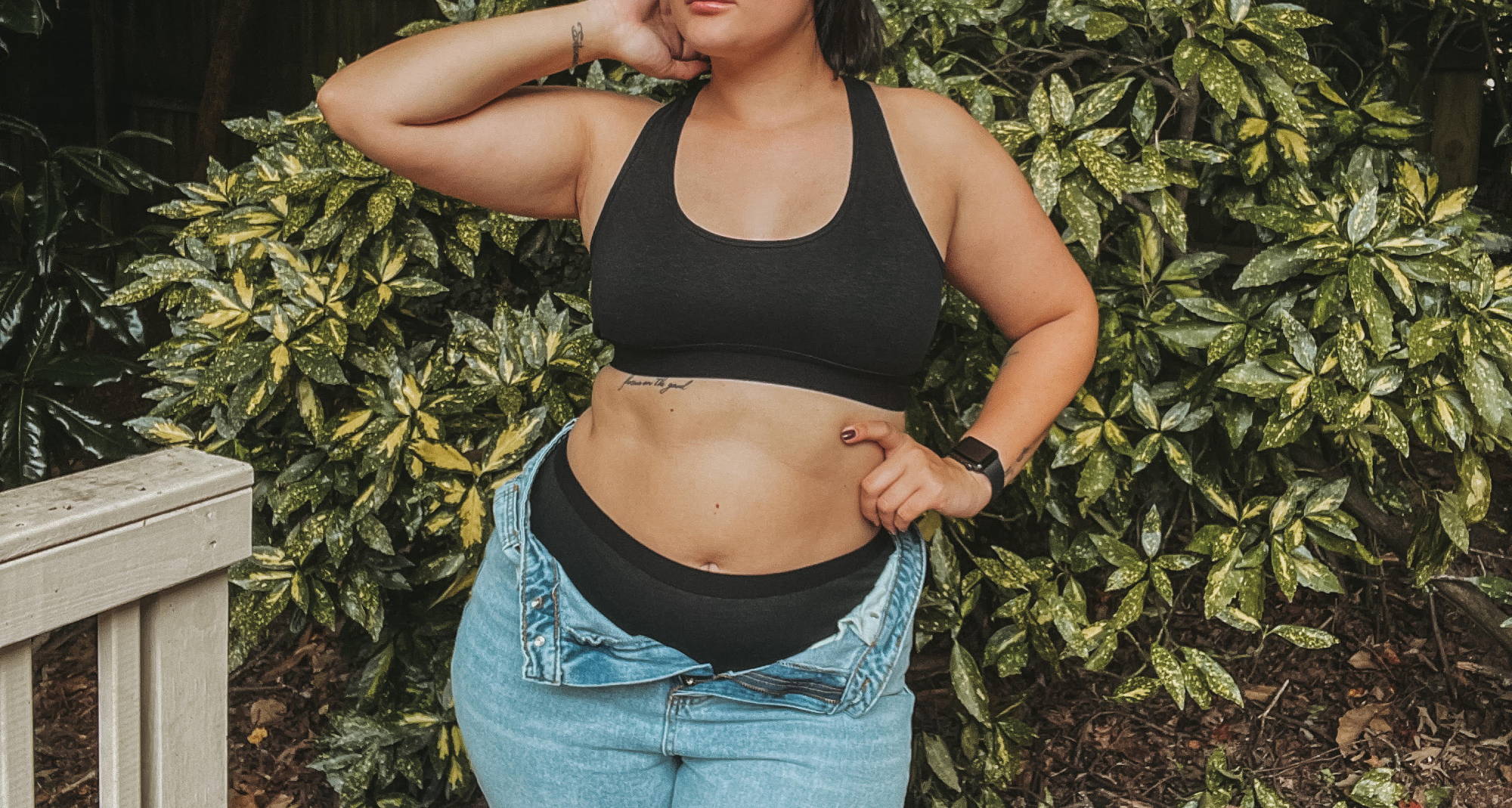 Woman in black bra, black underwear, and unbuttoned jeans standing in front of a bush. 