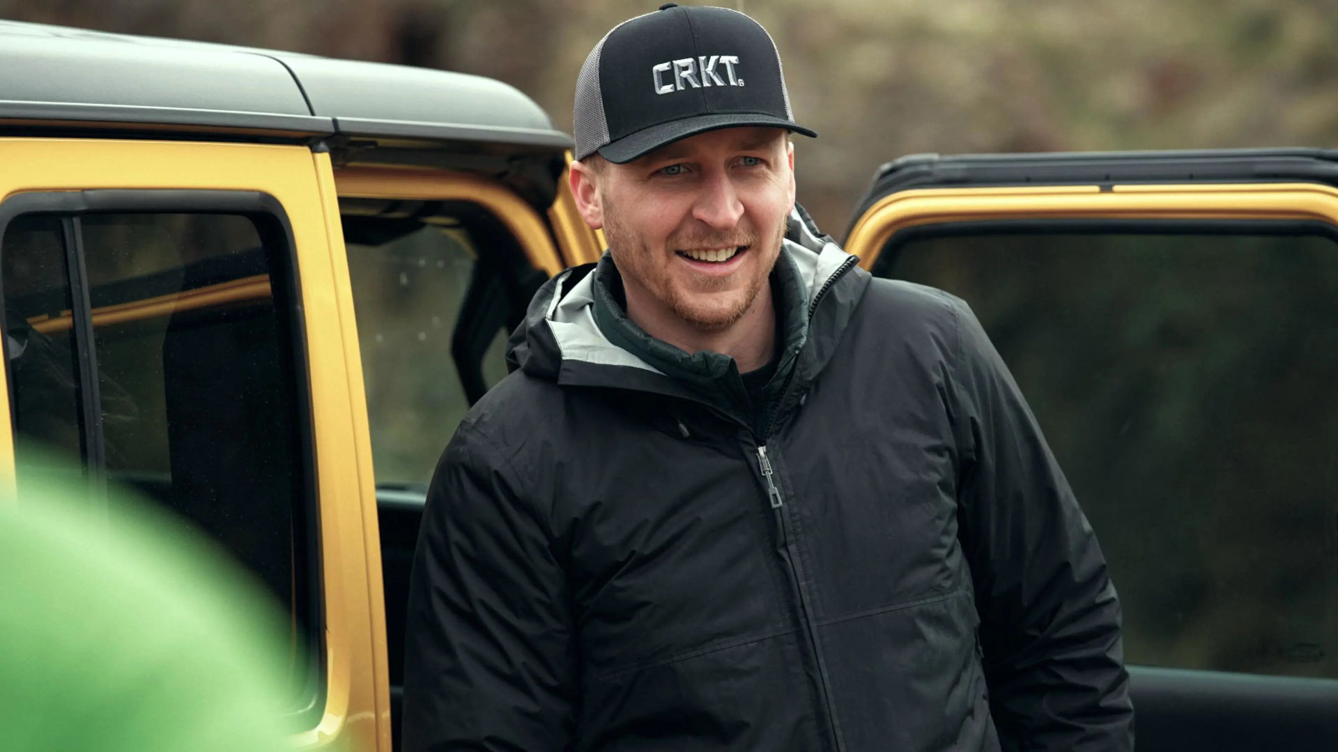 Man smiling near a yellow vehicle, wearing a black jacket and cap.