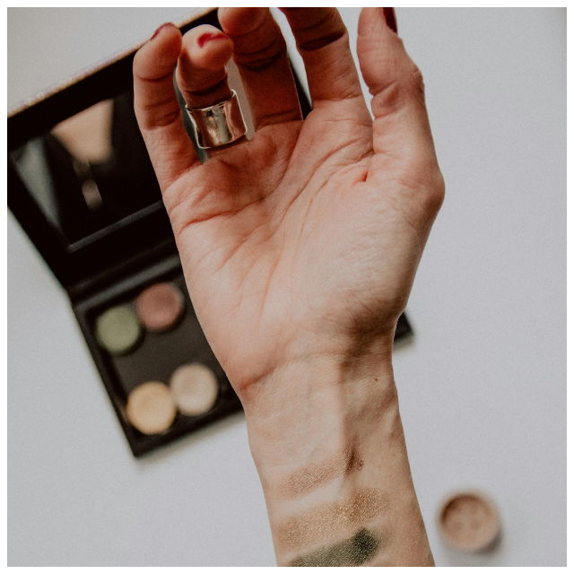 A hand in front of a makeup kit, with several test swatches of makeup on the wrist
