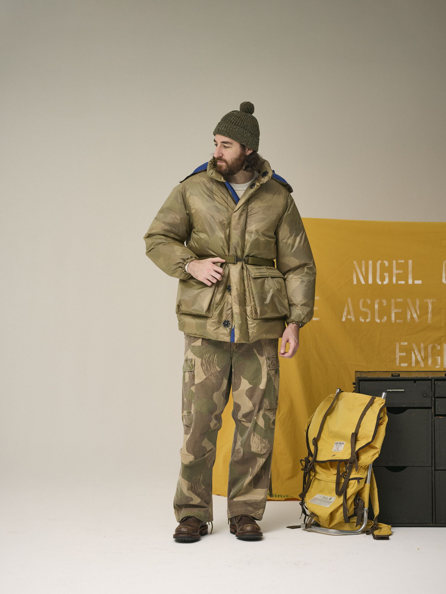 A man in a winter coat stands next to a yellow banner and backpack.