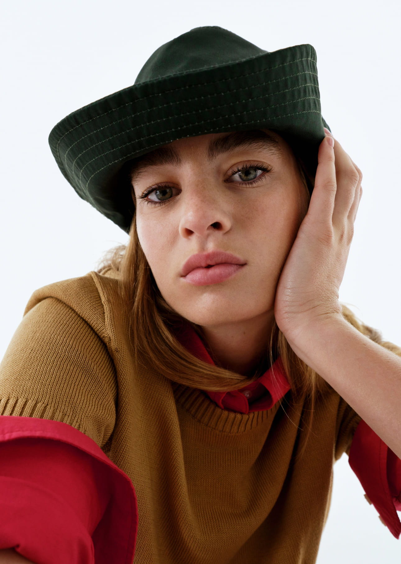 A woman wearing a green hat and a brown sweater, resting her chin on her hand.