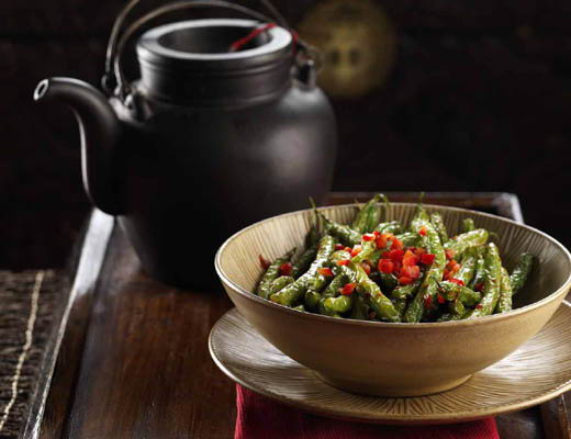 A bowl of green beans topped with red peppers, next to a teapot.