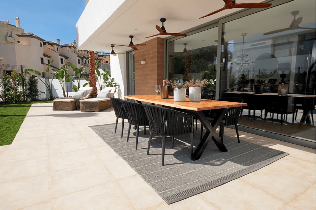 Gray outdoor dining rug placed beneath a rectangular wood table and black patio dining chairs, showing proper outdoor rug placement for a dining area on a tiled patio.