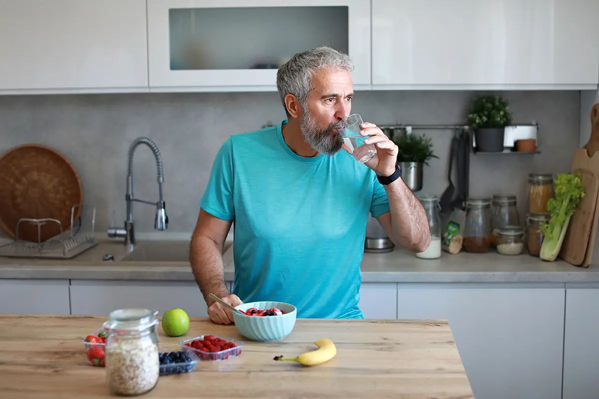 A man in a blue shirt drinks water in a kitchen.