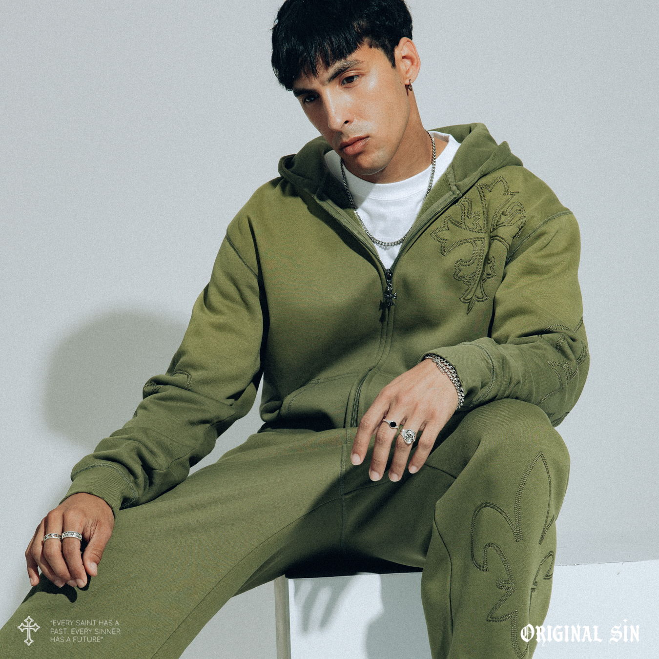 Young man in green hoodie and pants, seated against a light background.