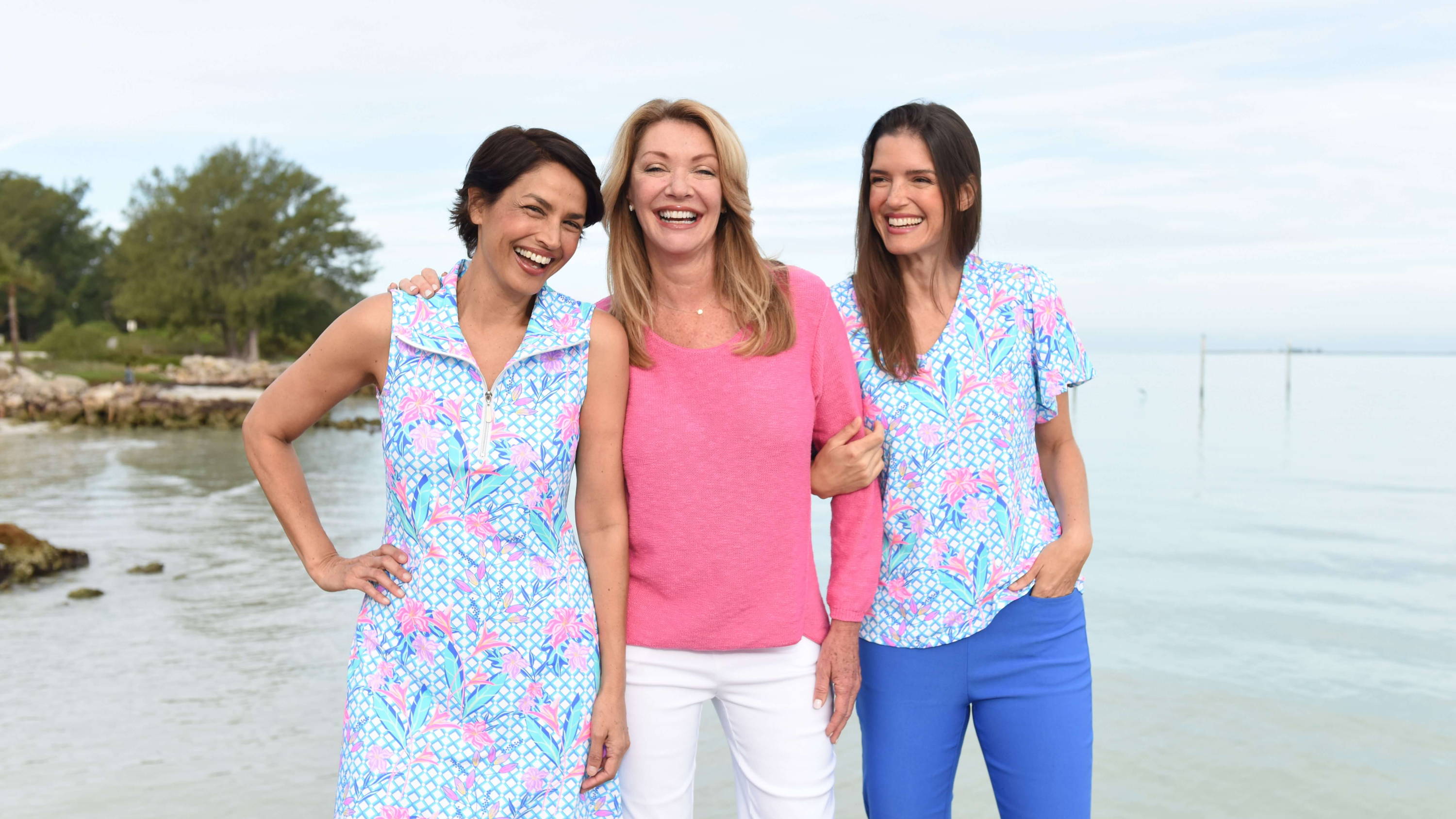 Three smiling women stand in front of a harbor. 
