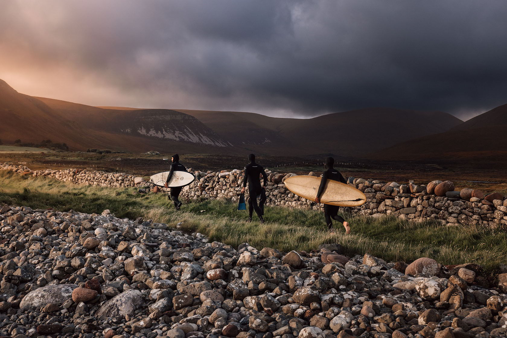 Surfers walking on a rocky shore under a dramatic sky.