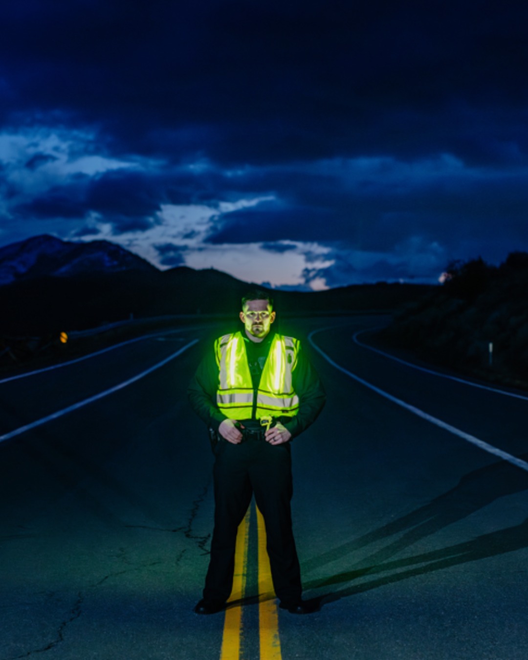 Person in reflective vest standing on a dark road with mountains in background.