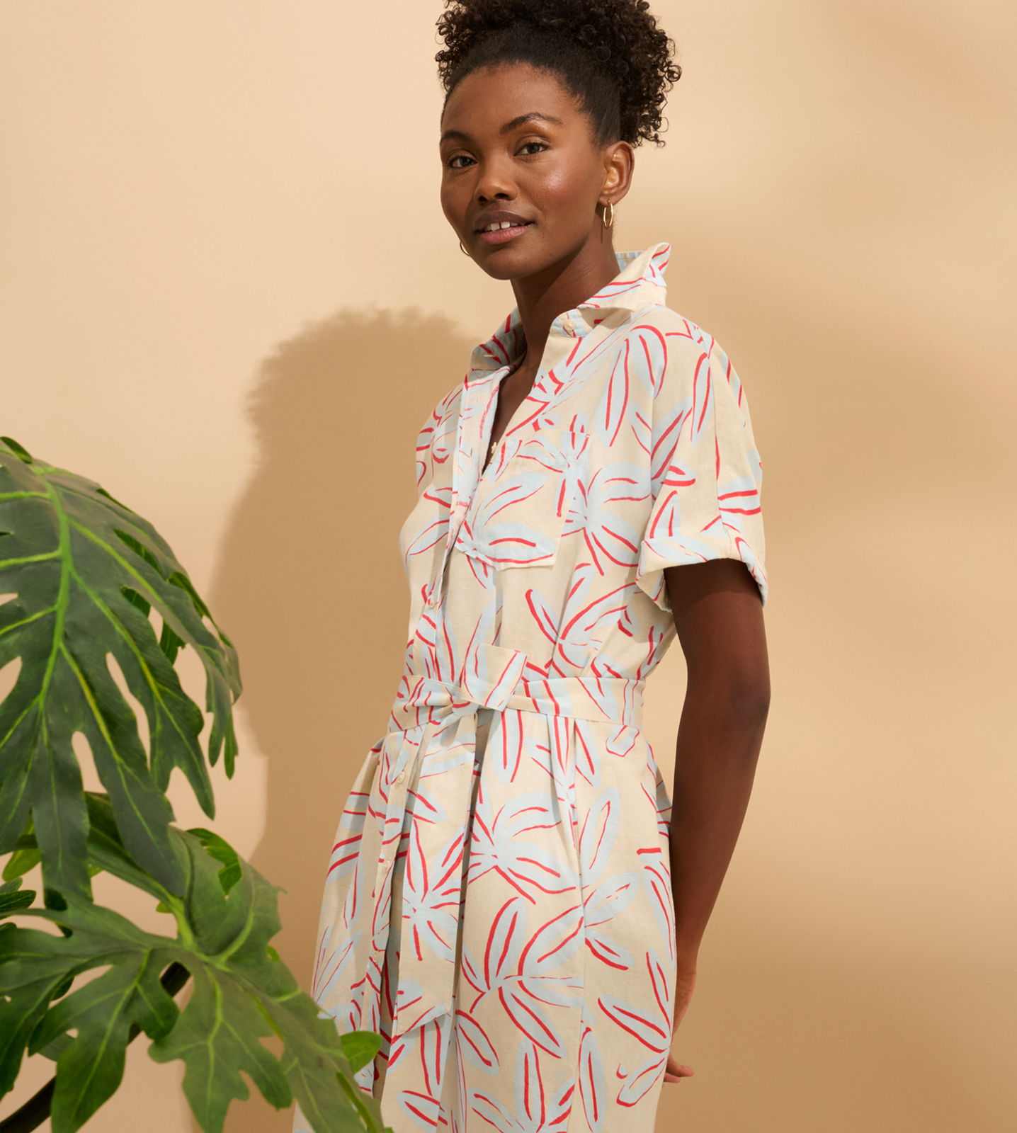 A woman in a patterned dress stands beside a plant against a beige background.