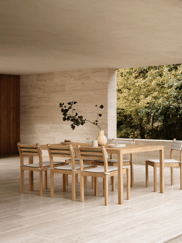 Covered outdoor dining area with a teak dining table and chairs set against light stone walls in a modern architectural patio.