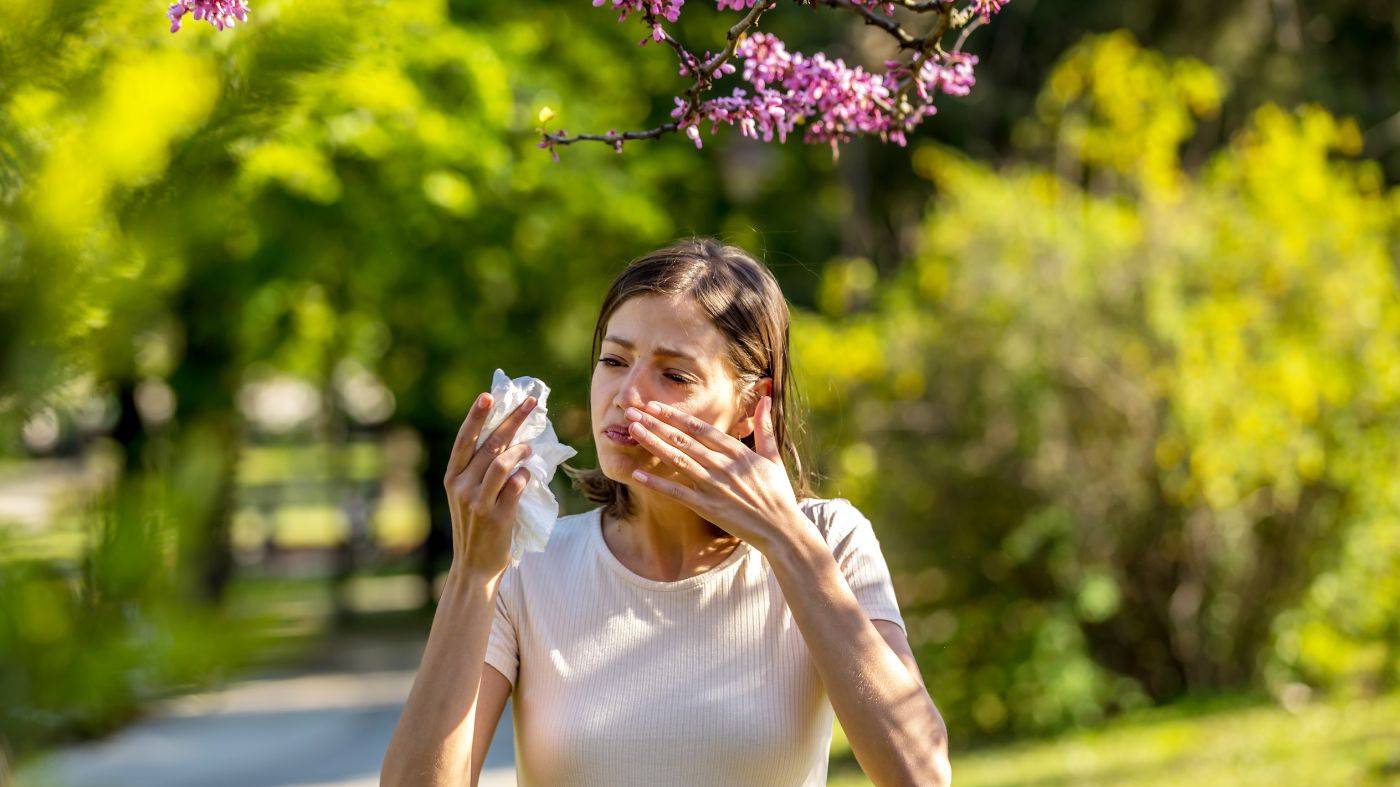 Woman blowing her nose in the park, surrounded by trees and flowers