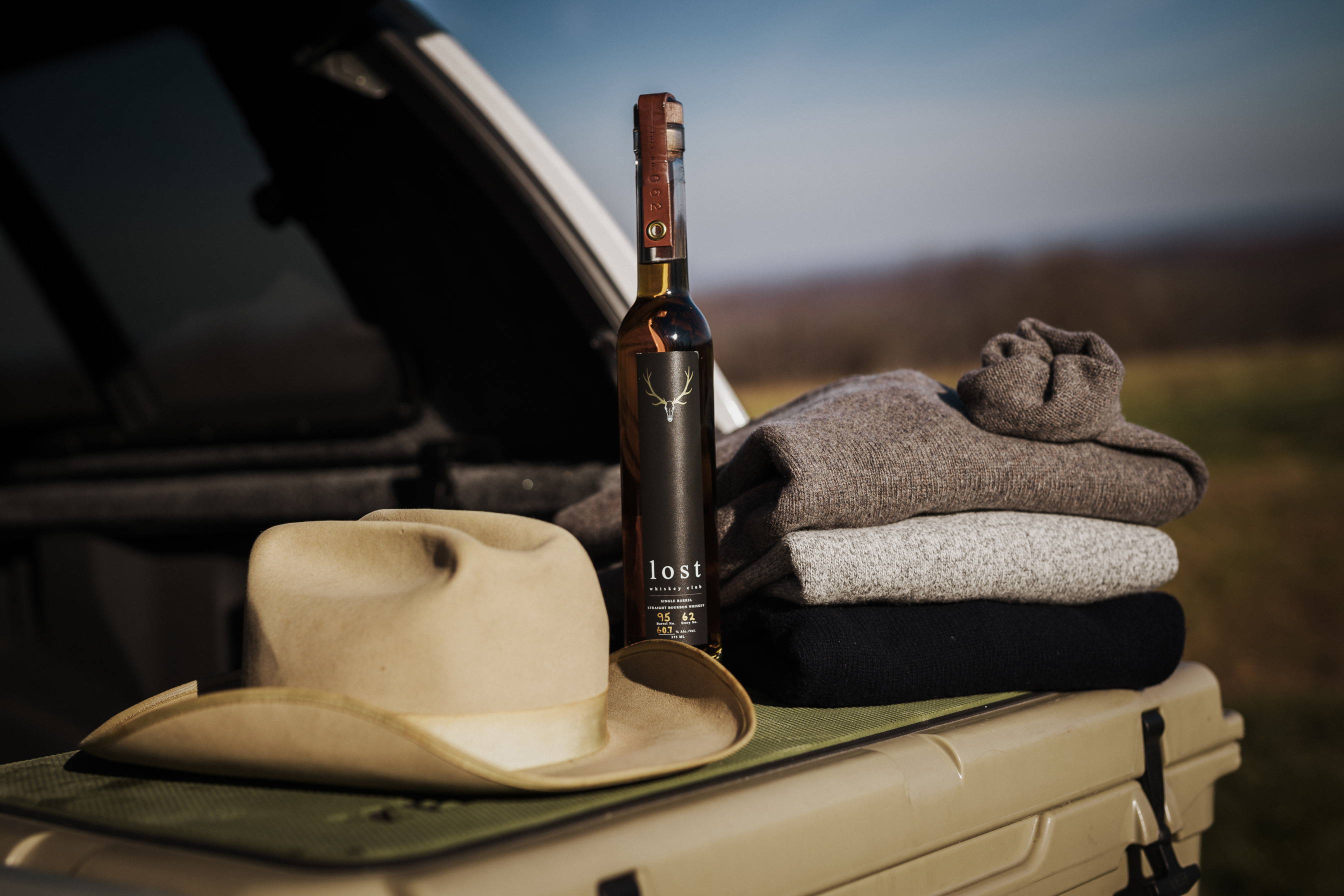 A detail shot of a tan hat, a Lost Whiskey bottle, and three Ledbury sweaters on top of a Yeti cooler.