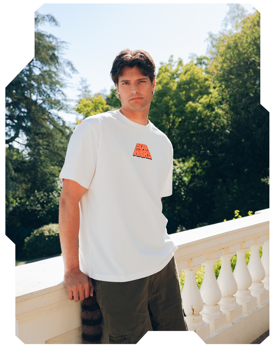 Man in white t-shirt standing by a balcony with greenery in the background.