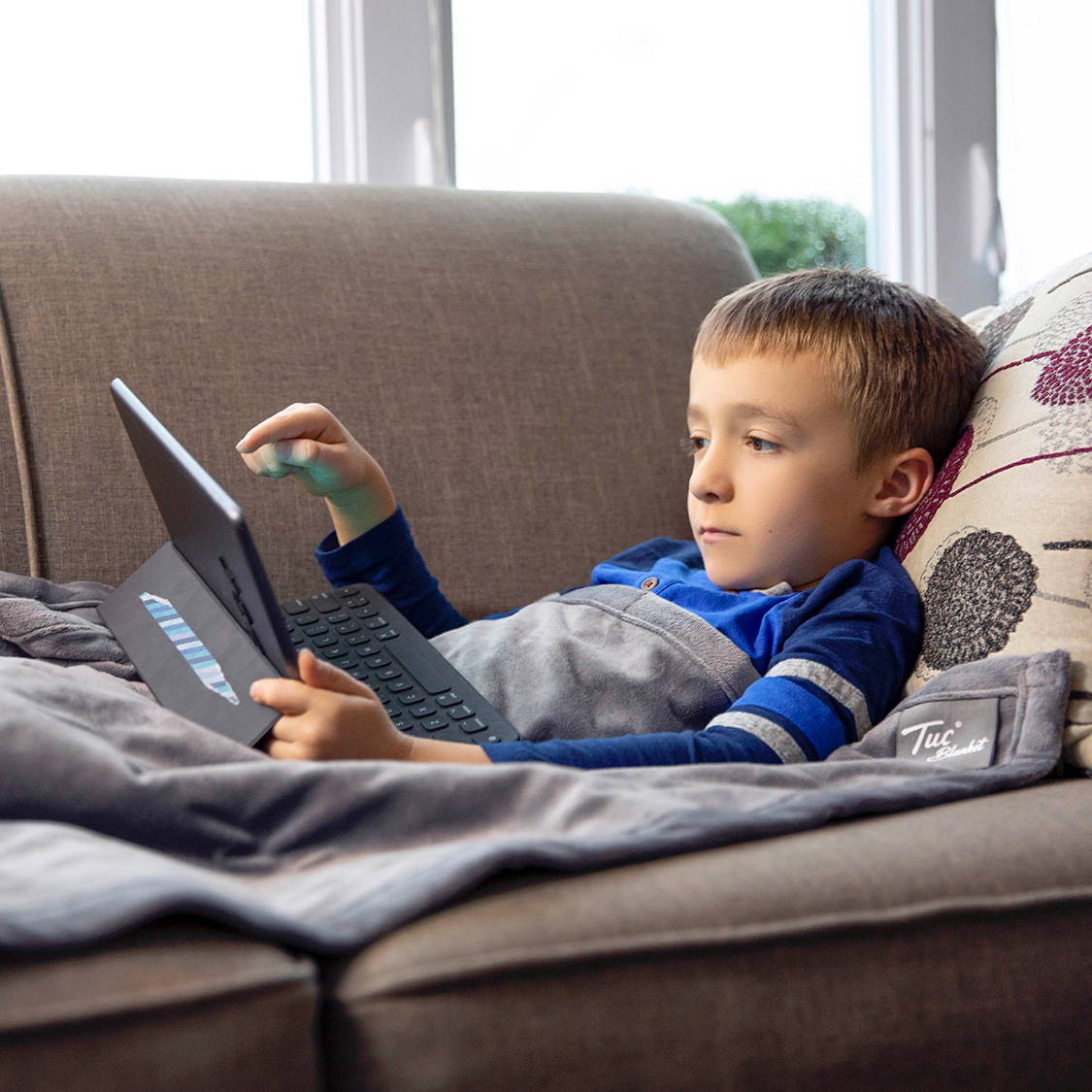 Boy relaxes on couch with tablet and Tuc Blanket.