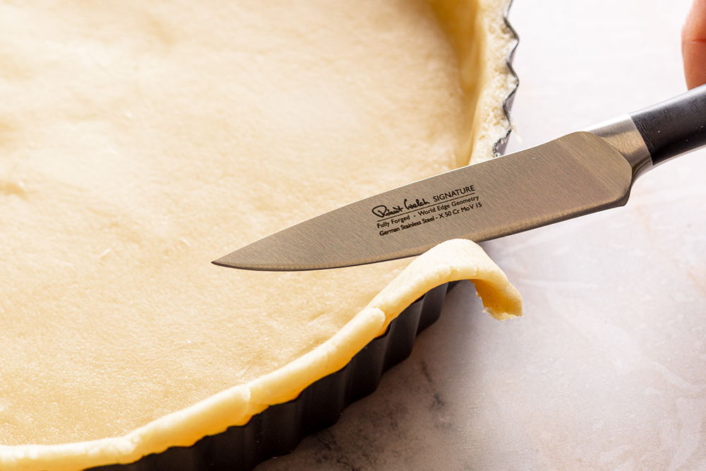A knife cutting the edge of unbaked pie crust in a tart pan.