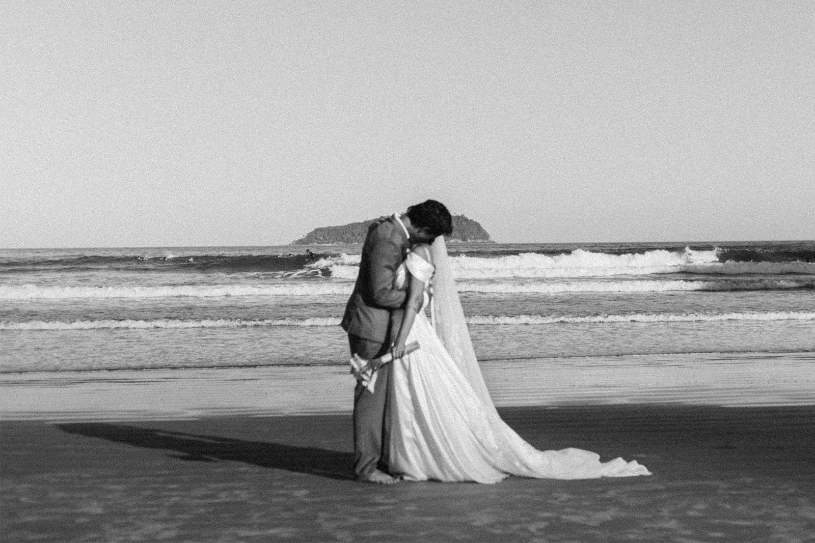 Couple kissing on the beach, dressed in wedding attire, black and white photo.