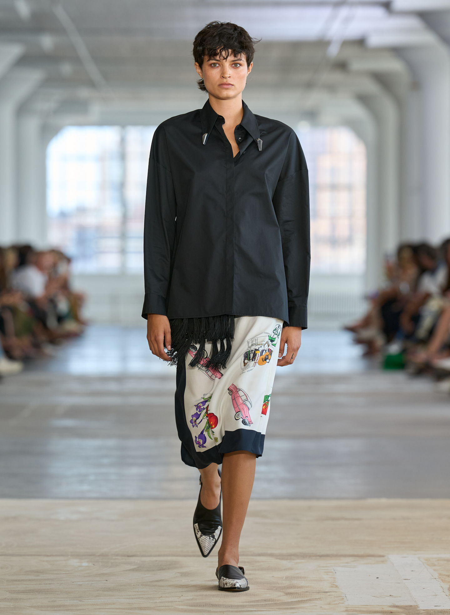 Model walking down runway in stylish outfit featuring black top and patterned skirt.