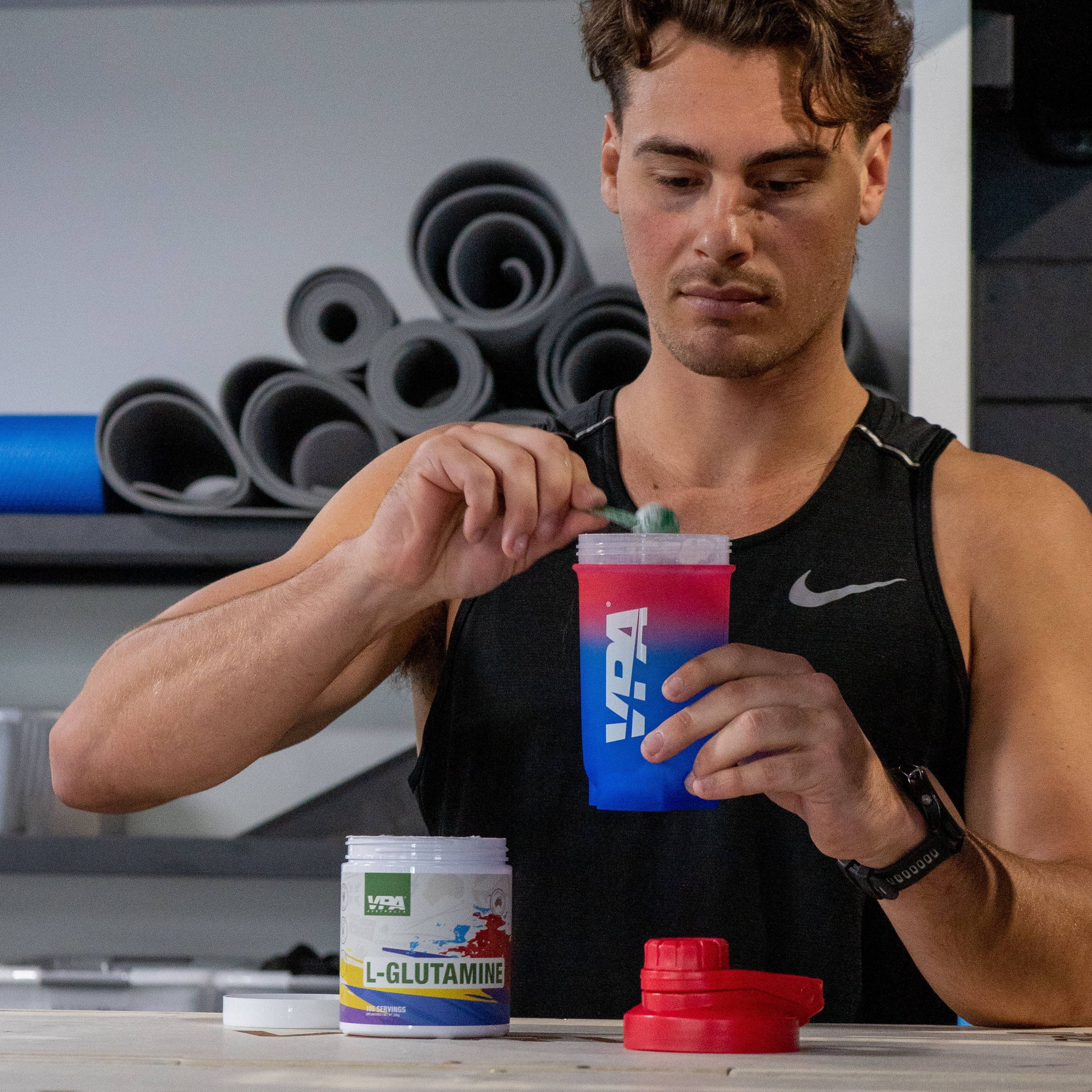 Man mixing l-glutamine powder in a red and blue gradient shaker at a fitness studio.