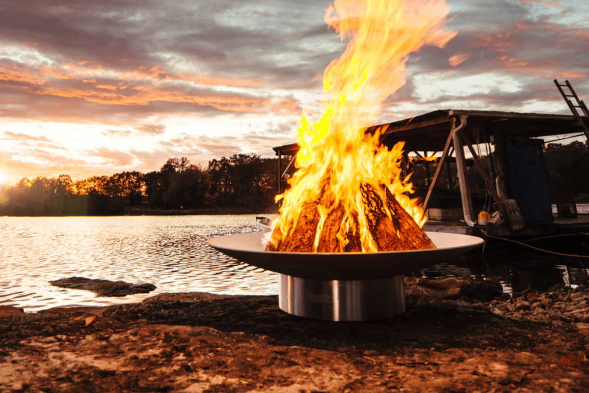 Logs burn with high flames from a stainless steel fire bowl on the bank of a lake.