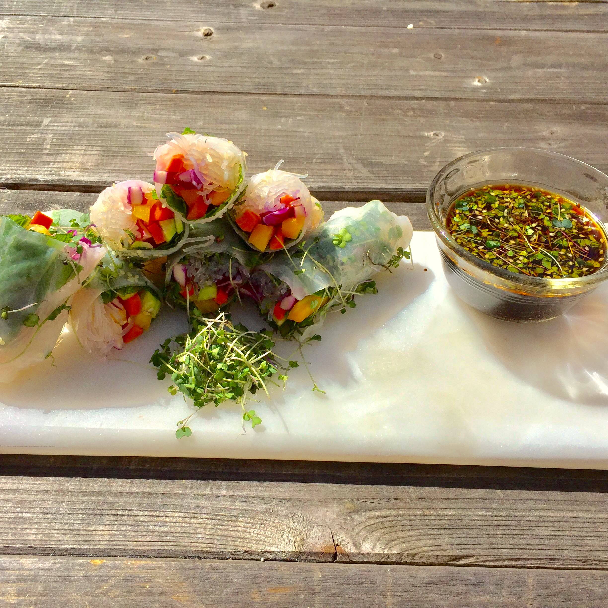 A stack of spring rolls made with rice paper, rice noodles, cabbage, carrots, cucumbers, bell peppers, and salad microgreens with a tamari, vinegar, and sesame oil dipping sauce.