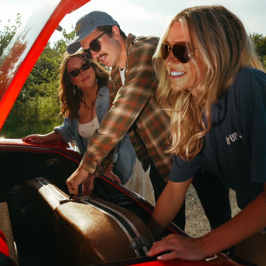 Three people loading a suitcase into a red car under sunlight.