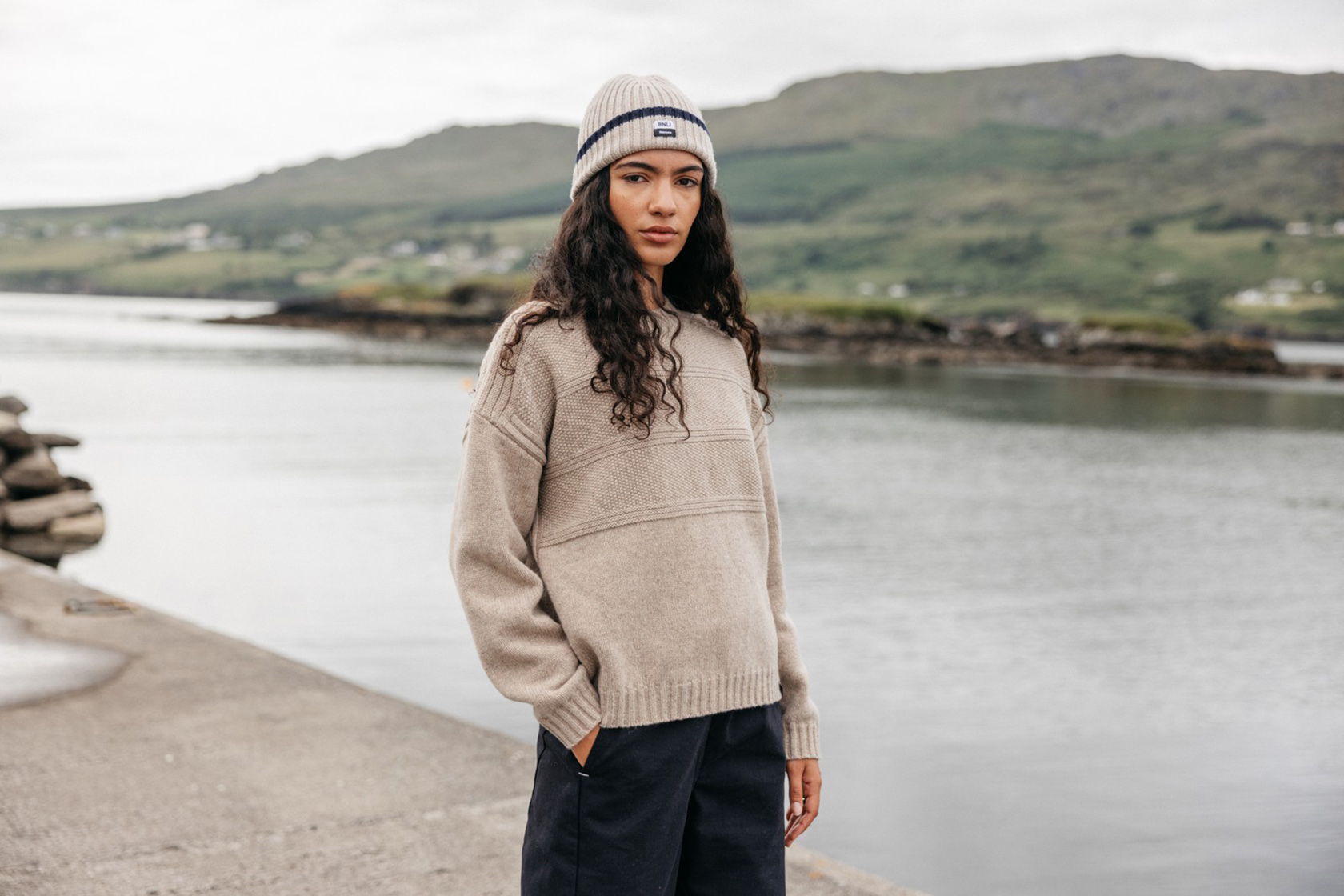 A woman in a beige sweater and hat near a body of water.