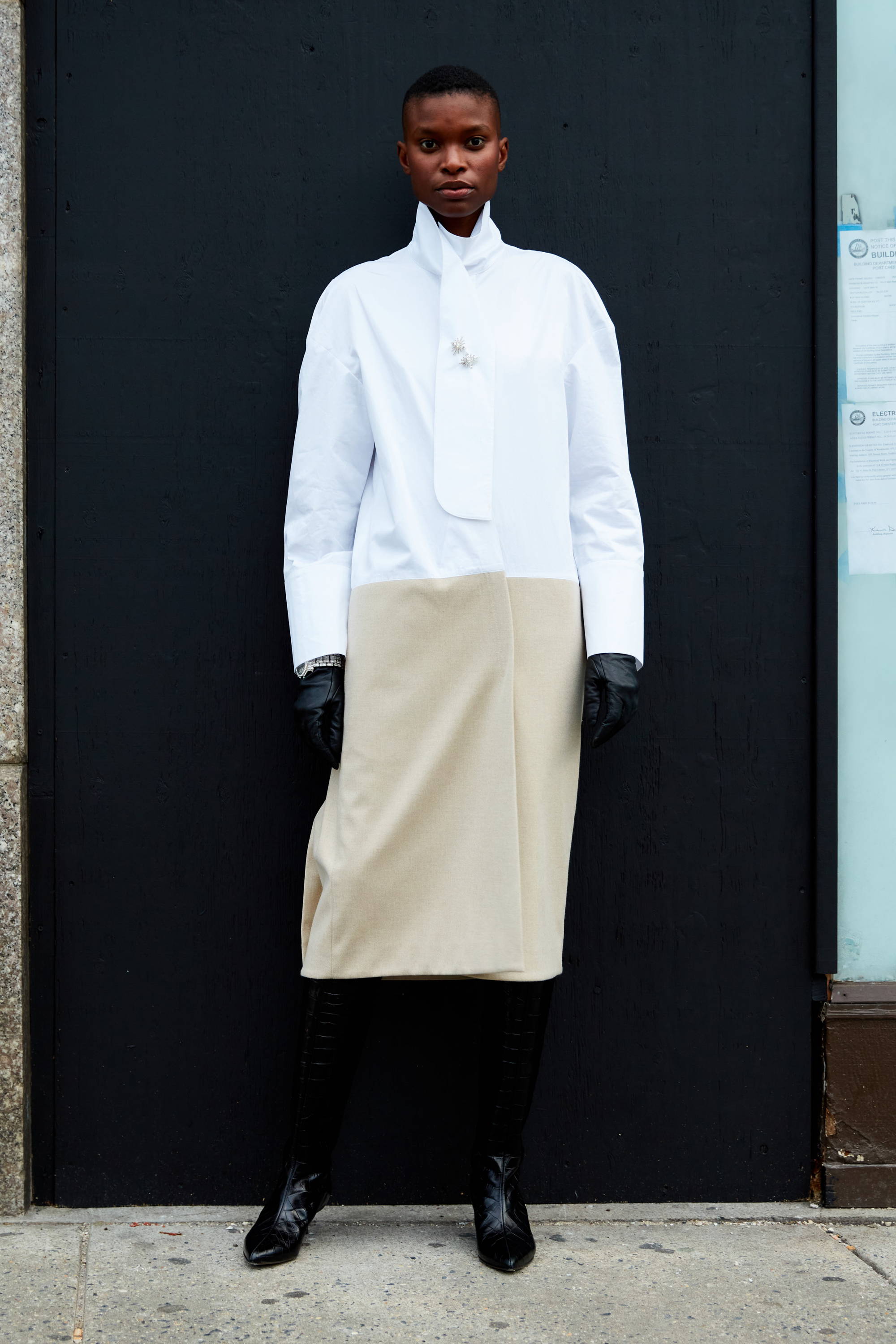 Person wearing a white shirt and beige skirt, standing against a black wall.