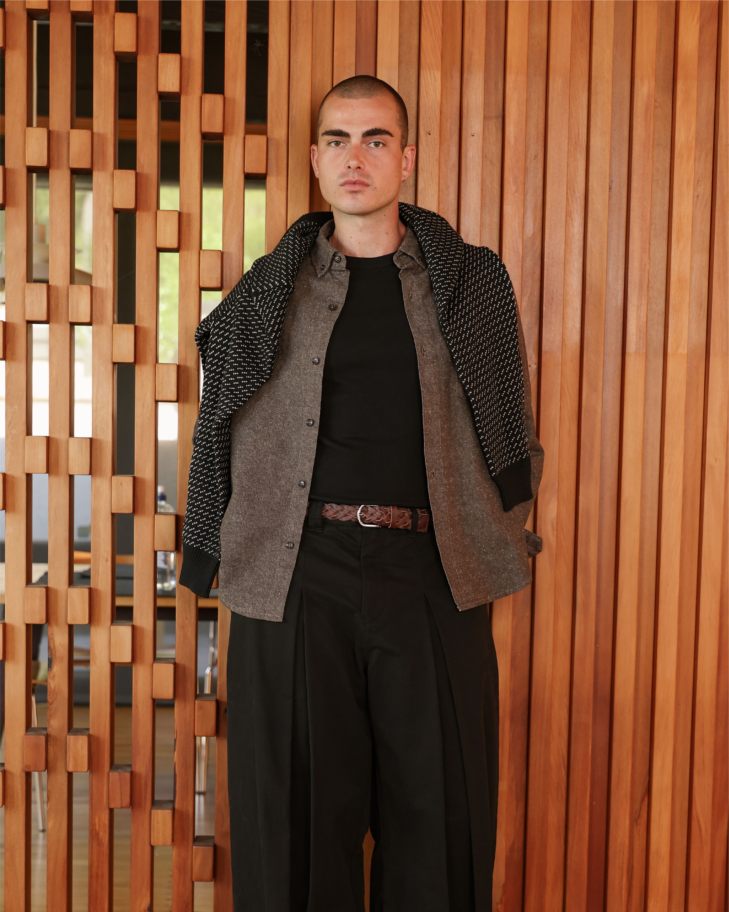 A man stands against a wooden wall, wearing casual fashion attire.