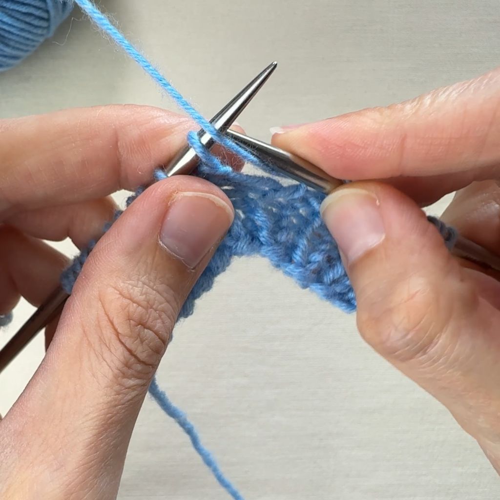 Detailfoto, wie eine linke Masche gestrickt wird. Im ersten Schritt kommt der Faden vor die Arbeit.