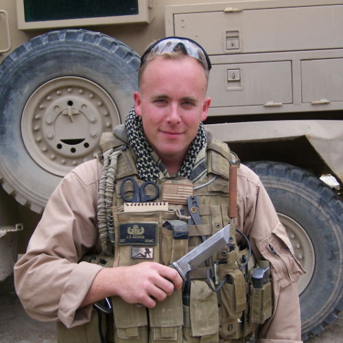Soldier in tactical gear holding a firearm, standing near military vehicle.