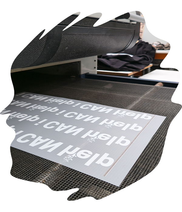 Sheets of cured DTF transfers showing mirrored white text and logo designs covered in adhesive powder sittong on the conveyor belt of the oven