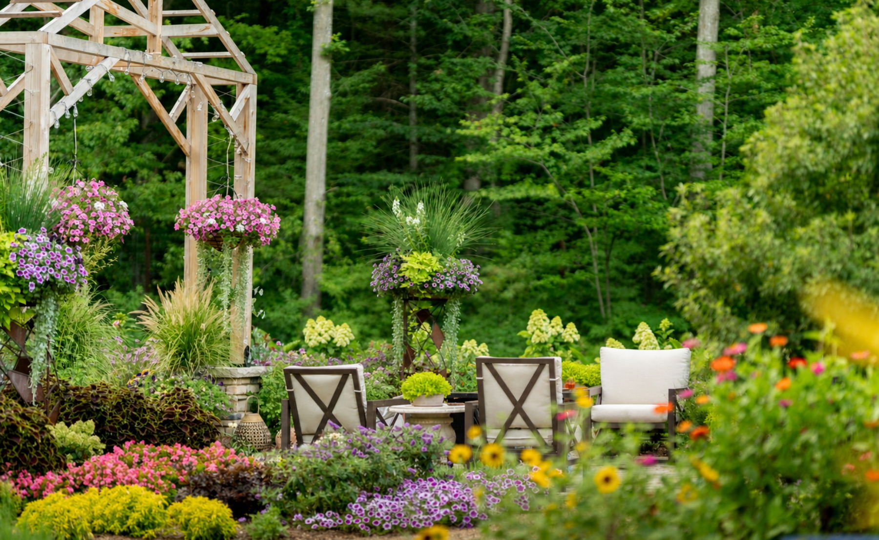 Bright, lush garden patio with a wooden pergola, colorful flowering plants, and empty outdoor seating surrounded by greenery and trees.