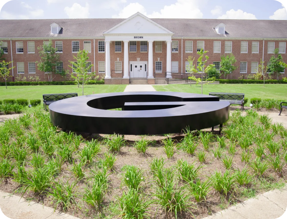 Black circular sculpture in front of a brick building with greenery.