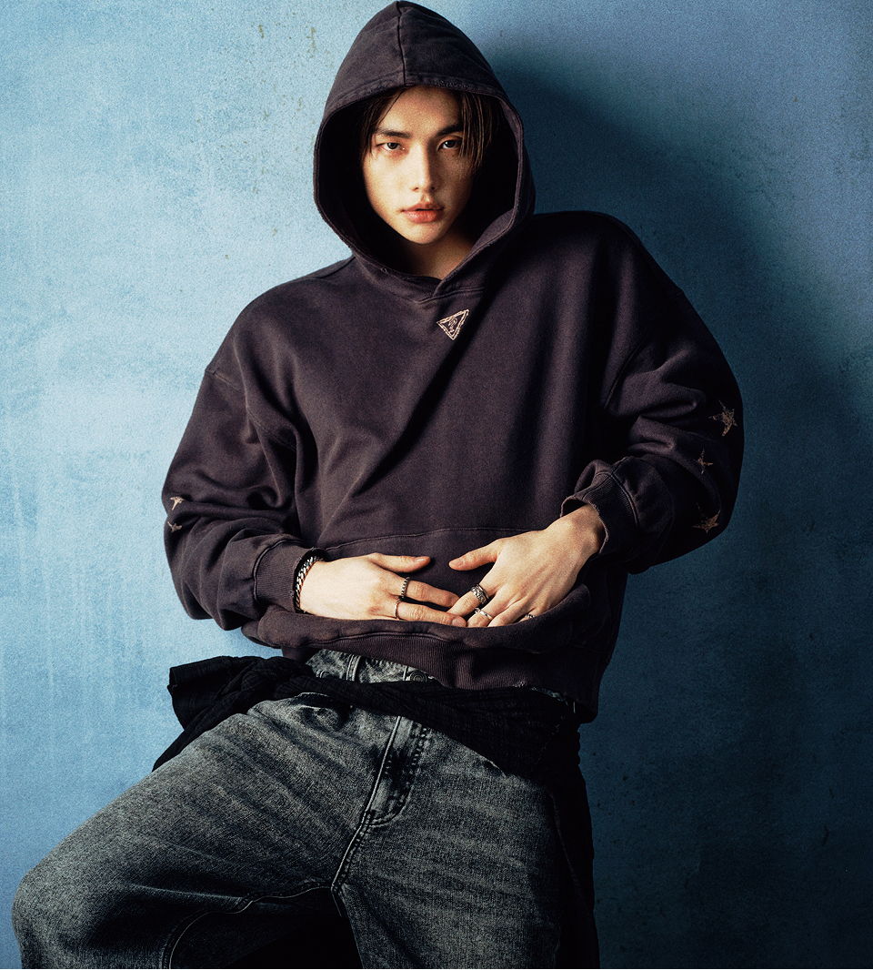 Hyunjin in a new Hyunjin x GUESS hoodie posing against a blue background.