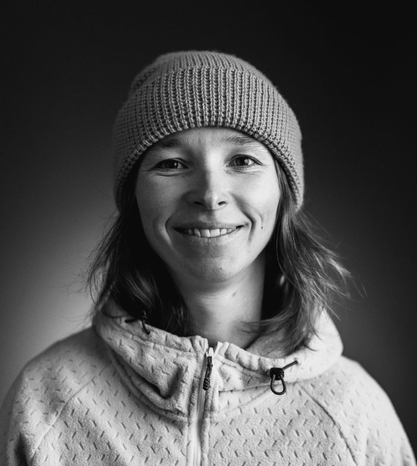 Smiling person wearing a beanie and hoodie against a dark background.
