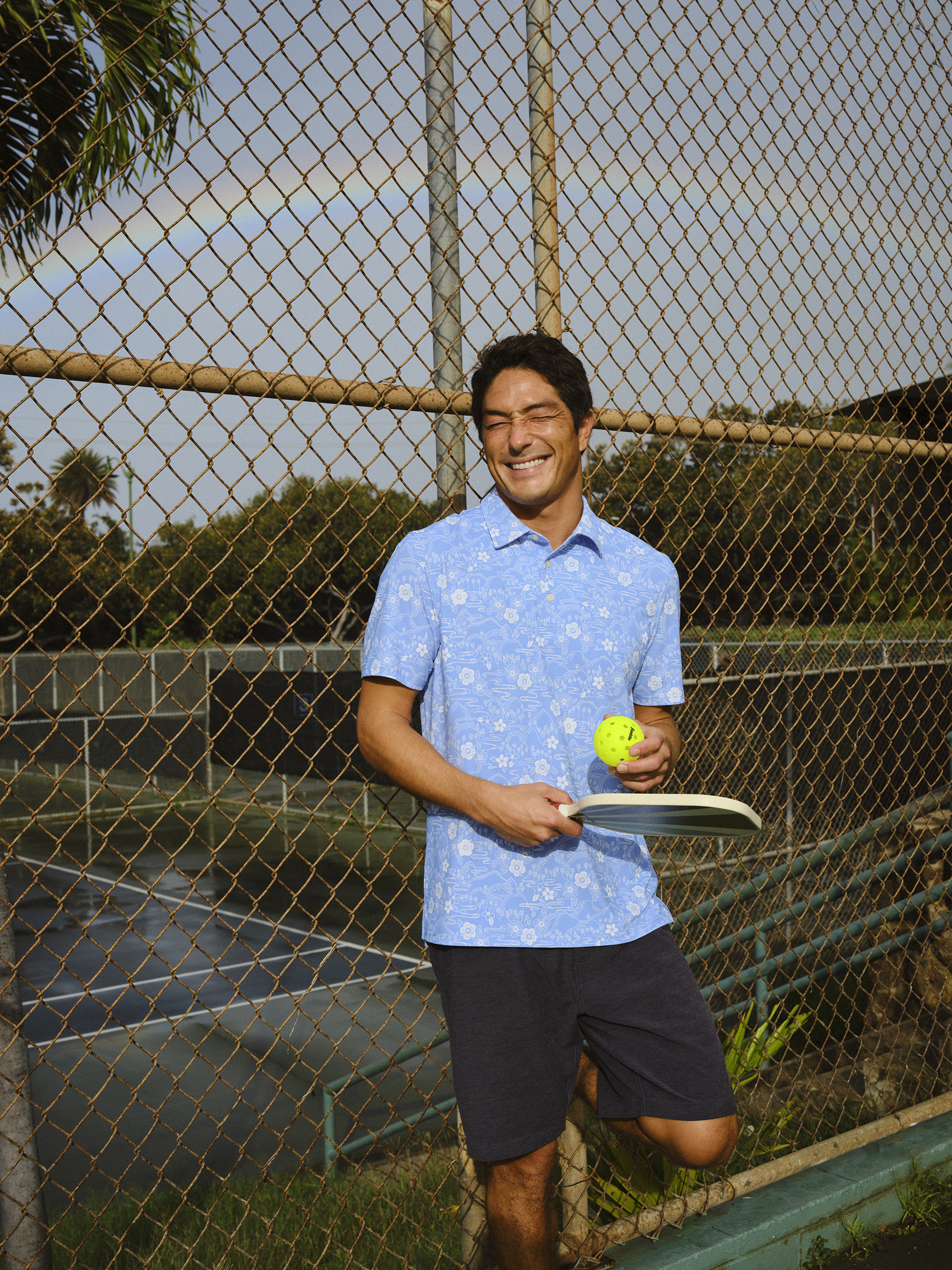 Smiling man holding a tennis racket and ball near a fence.