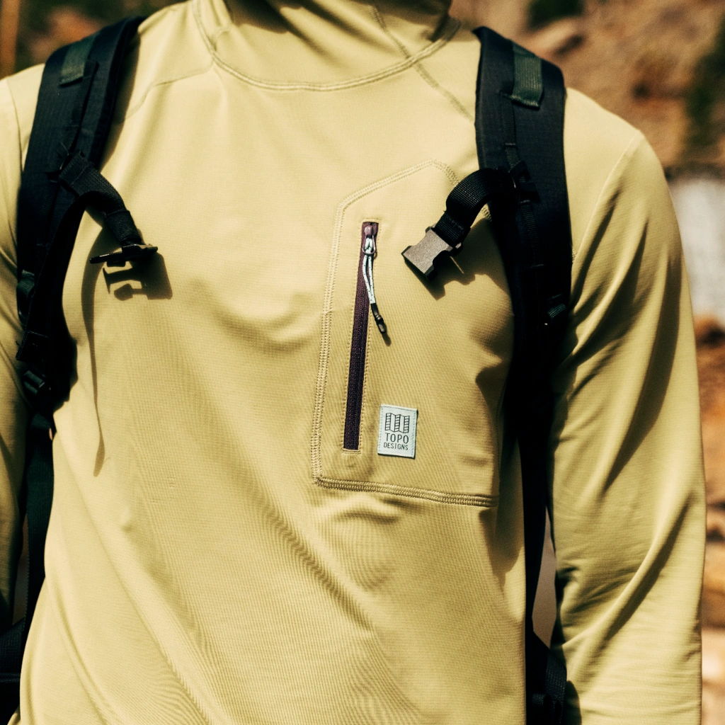 Person wearing a tan long-sleeve shirt with a zipper pocket and backpack.