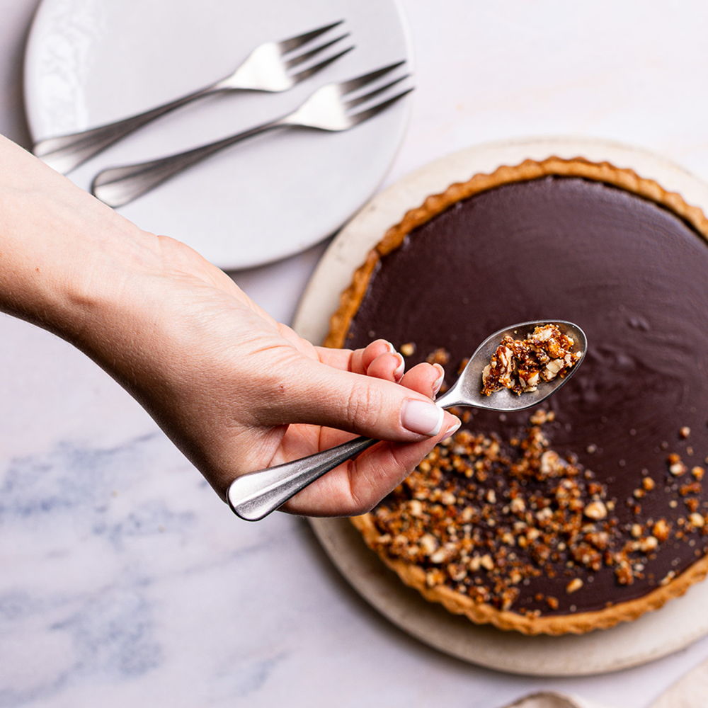 Hand holding a Baguette Bright spoon above a chocolate tart with nuts.