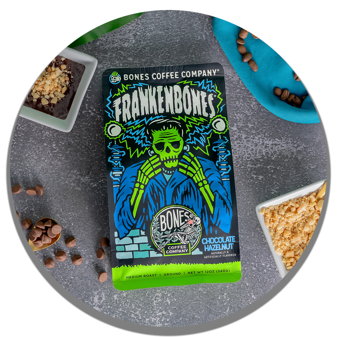 The front of a 12 ounce bag of Bones Coffee Company Frankenbones coffee. Its flavor is chocolate hazelnut, and it has a green skeleton with blue and green electricity scattered around on the art. It is on a table with chocolate and hazelnuts.